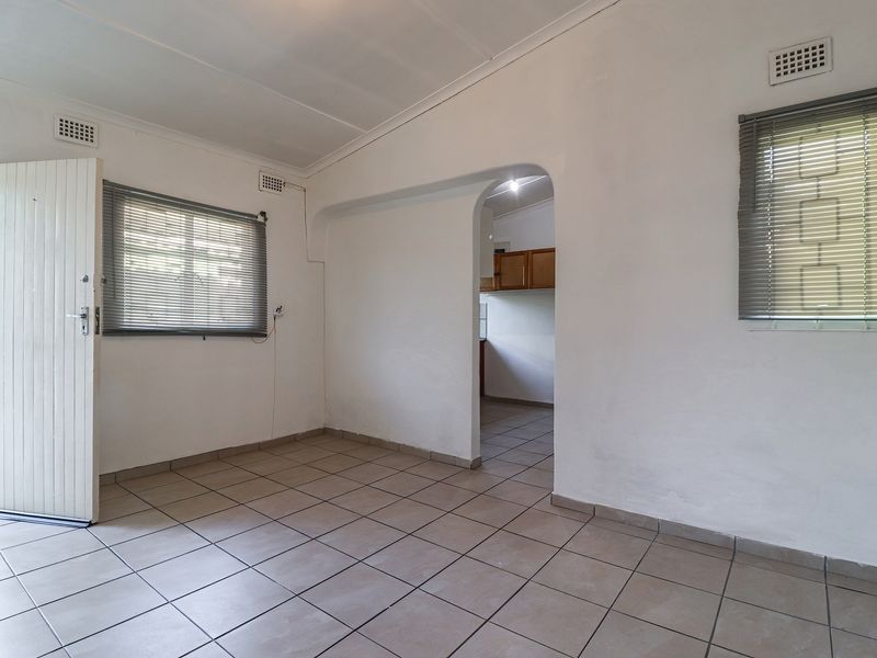 An empty living room featuring tiled flooring, an arched entryway leading to the kitchen, and natural light through windows with fitted venetian blinds.