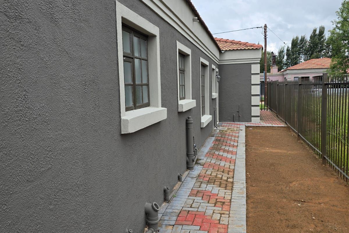 Side exterior view of the house featuring a paved walkway, textured grey facade, and metal perimeter fencing.