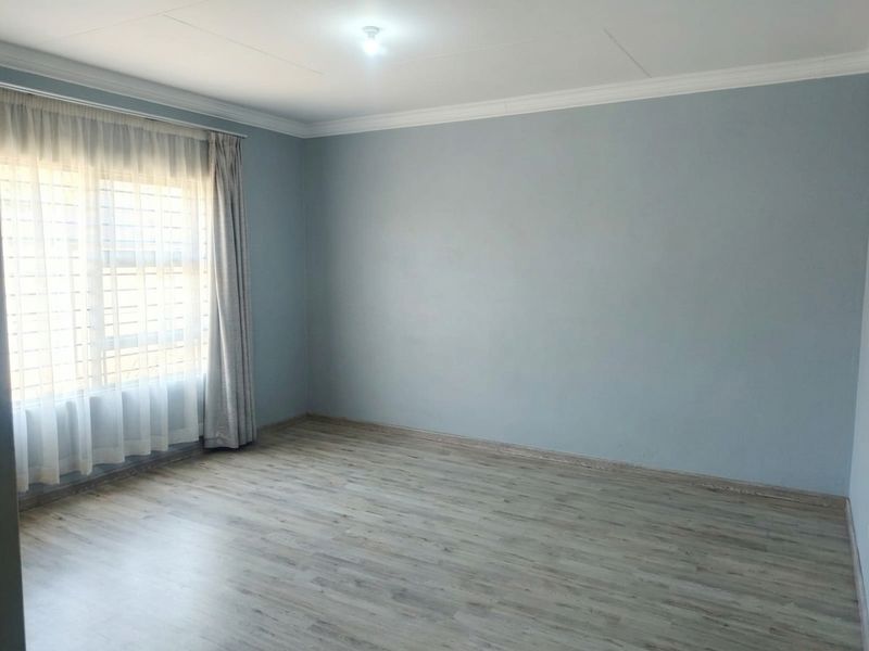 A bright, empty bedroom featuring light-toned laminate flooring, neutral blue walls, and window security bars.