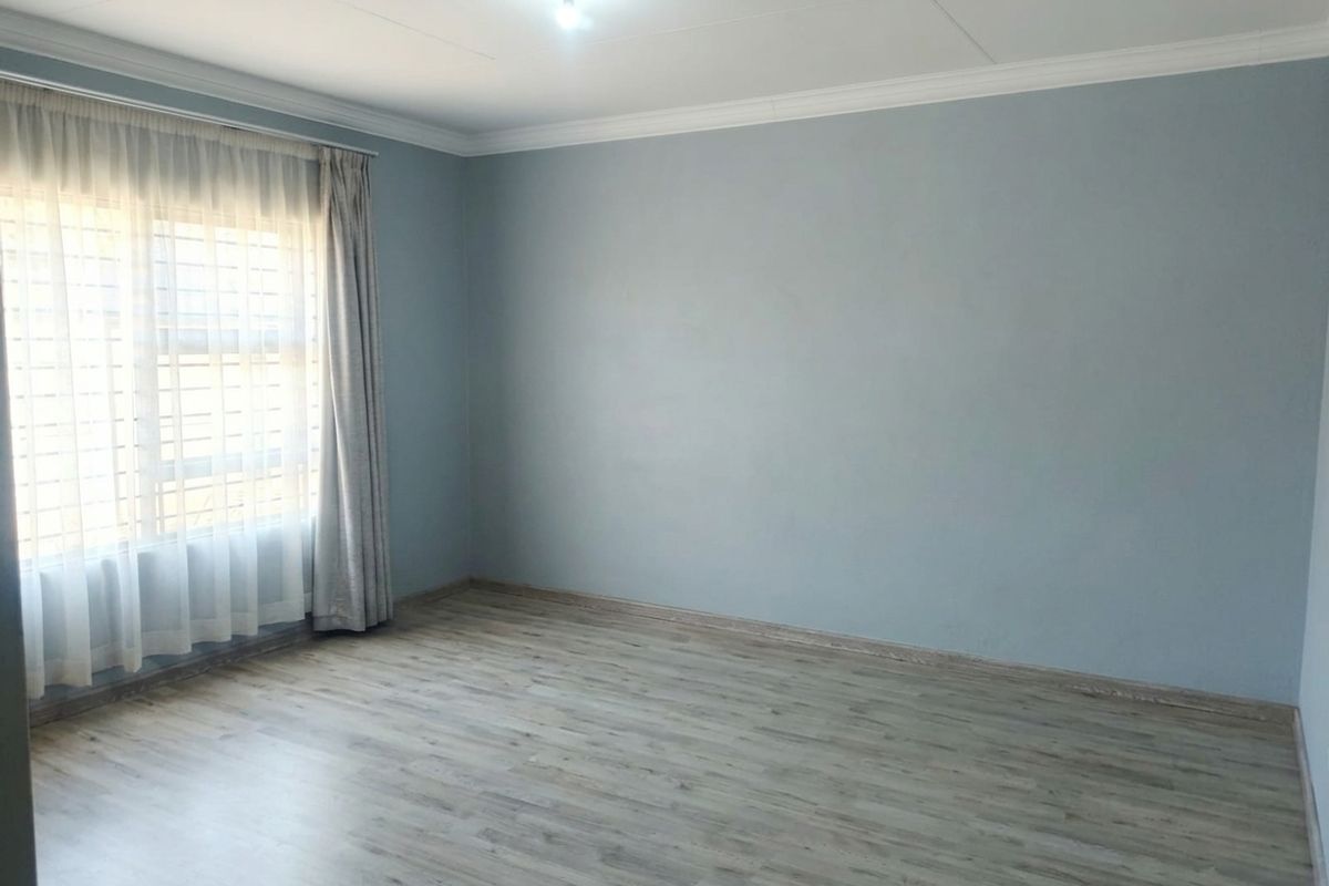 A bright, empty bedroom featuring light-toned laminate flooring, neutral blue walls, and window security bars.