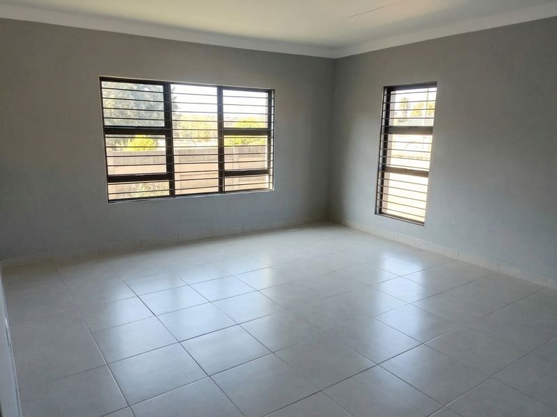 A bright, empty living area featuring neutral tiled flooring, grey walls, and large windows fitted with security bars.