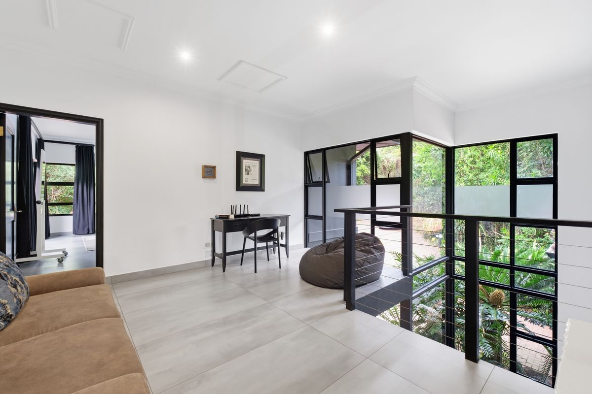 A bright, modern living area featuring large format floor tiles, black framed windows, and a sleek metal balustrade overlooking the garden.