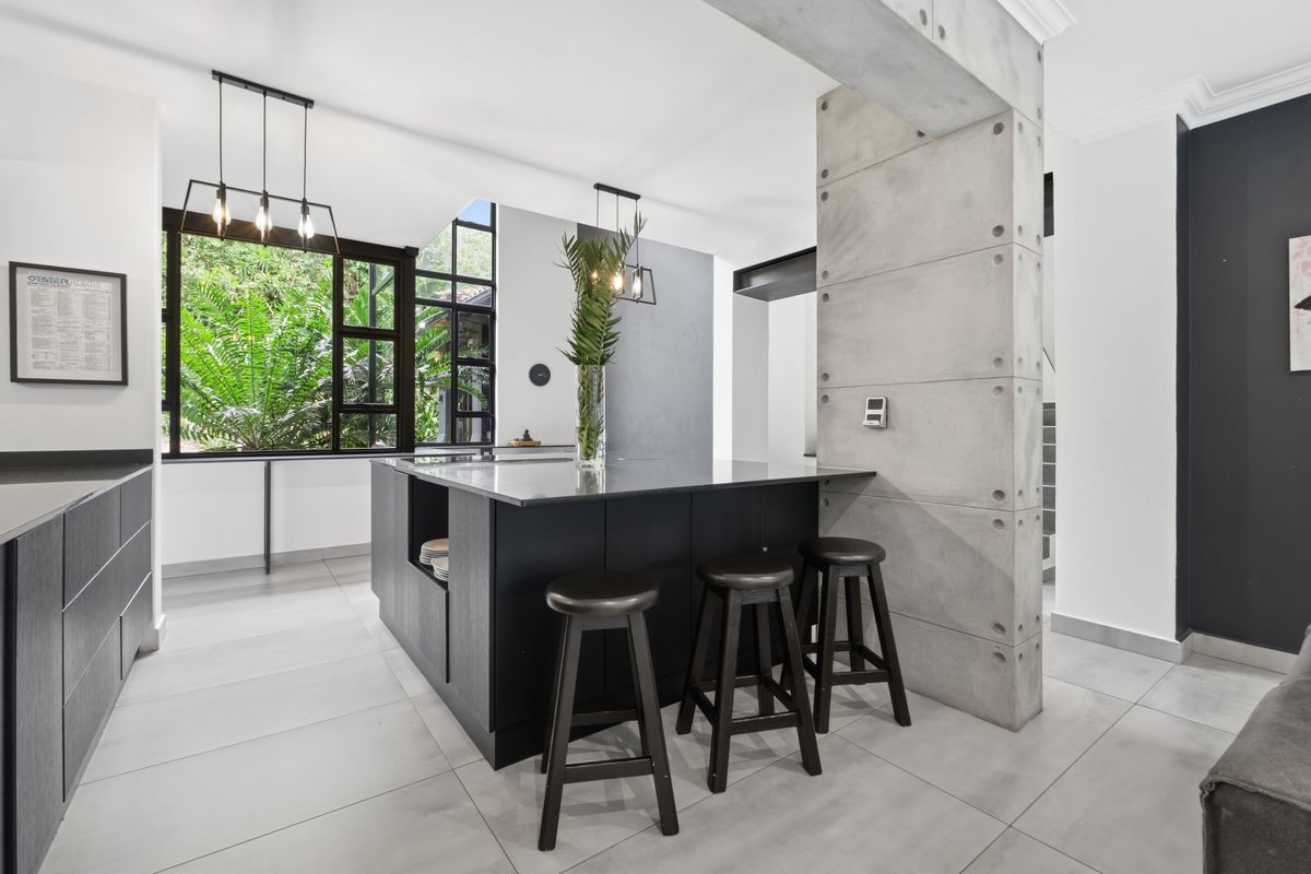 Modern kitchen featuring a central island with stone countertops, black cabinetry, and a striking exposed concrete pillar, complemented by large windows and tiled flooring.