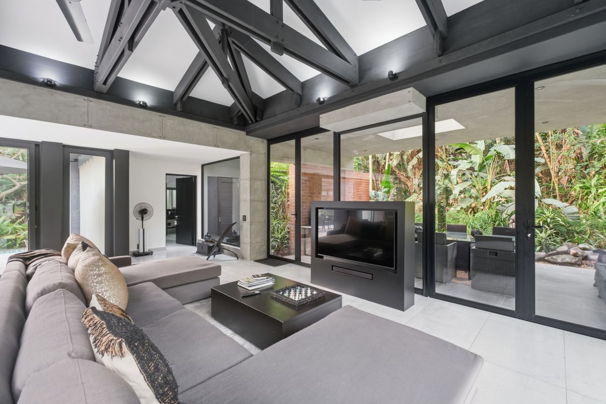 Modern living room featuring exposed dark timber beams, large format floor tiles, and expansive glass sliding doors opening to a lush garden view.