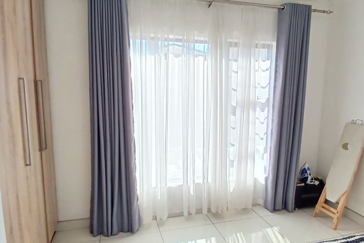 A bright bedroom featuring built-in wooden cabinetry and large windows dressed with floor-to-ceiling curtains, finished with neutral tiled flooring.