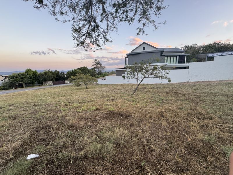 A vacant residential plot featuring natural grass coverage, boundary walling, and scenic views of the surrounding neighborhood and horizon.