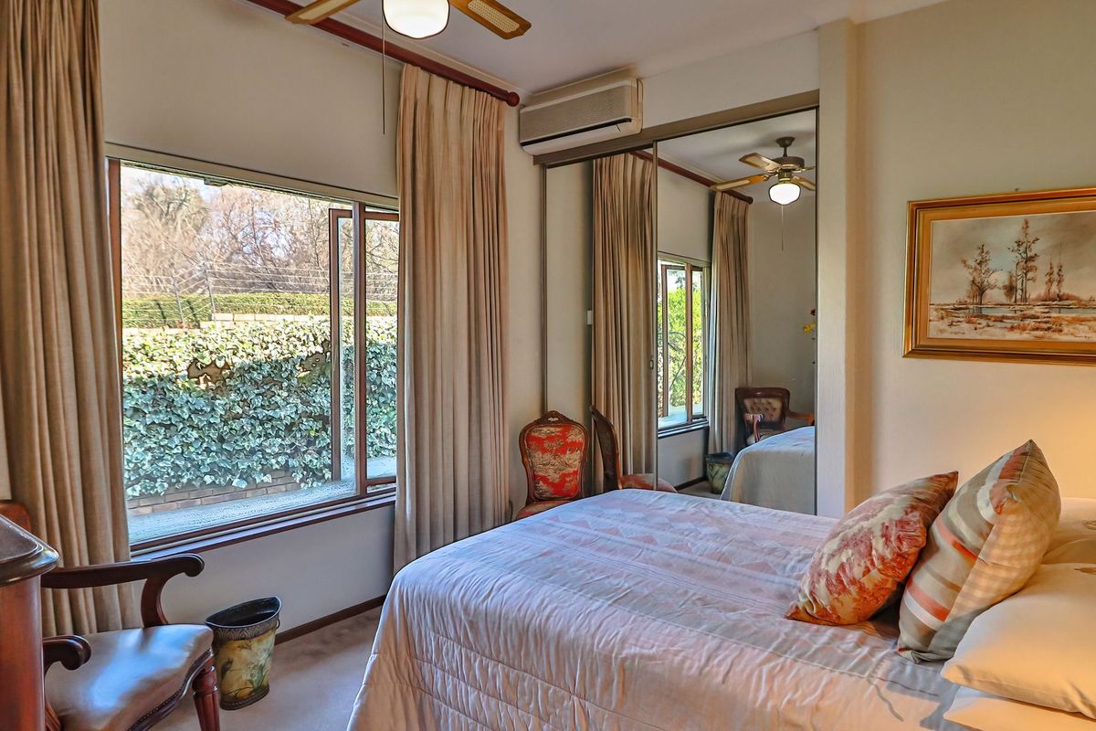 A bedroom featuring built-in mirrored wardrobes, a wall-mounted air conditioning unit, and a ceiling fan, with large windows overlooking the garden.
