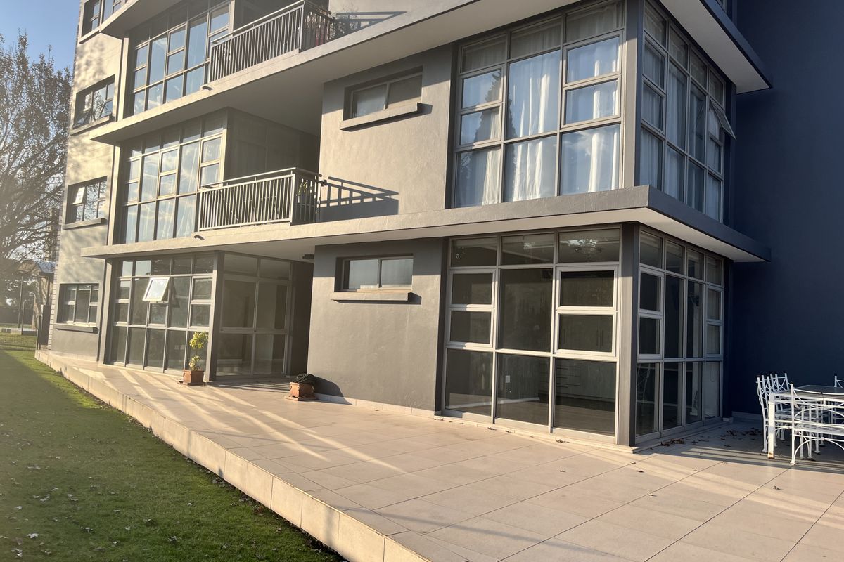 Exterior view of a modern apartment building featuring large glass windows, private balconies, and a spacious tiled patio area overlooking the garden.