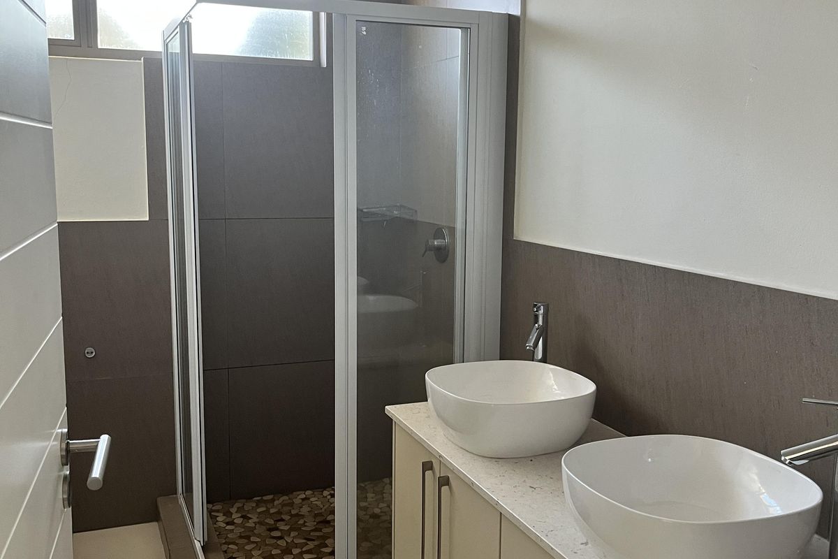 Modern bathroom featuring a double vanity with vessel sinks, a glass-enclosed shower with pebble-style flooring, and neutral wall tiling.