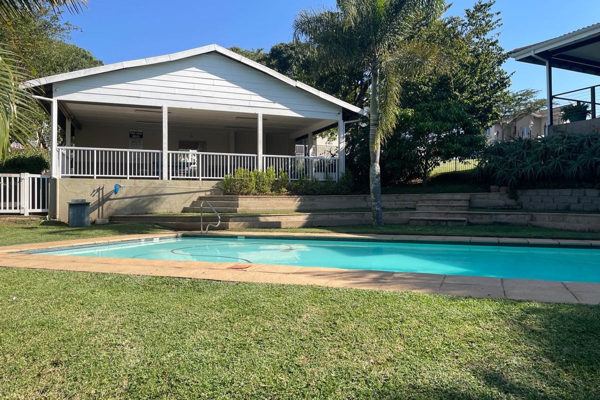 A well-maintained communal swimming pool area featuring a paved surround, tiered landscaping, and an adjacent covered patio structure.