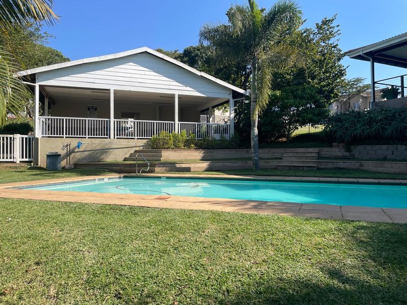 A well-maintained communal swimming pool area featuring a paved surround, tiered landscaping, and an adjacent covered patio structure.