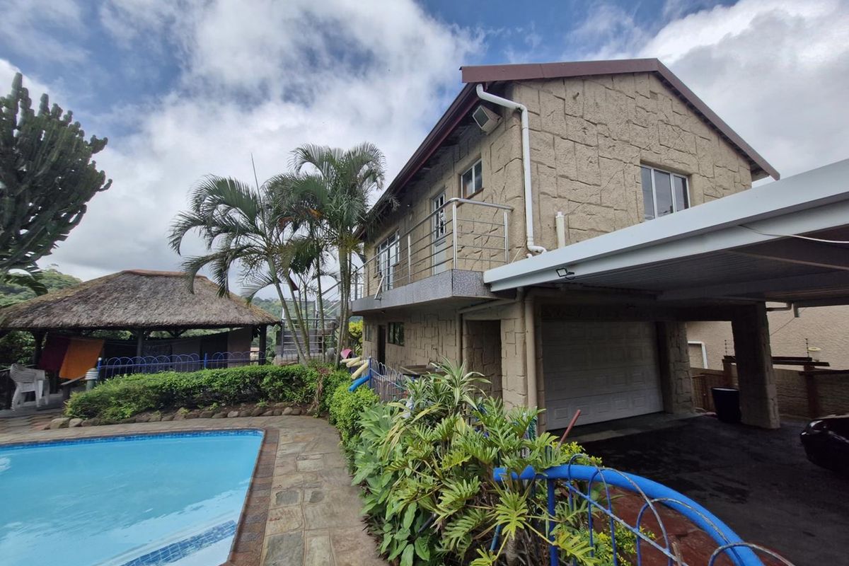 Rear exterior view featuring a swimming pool, stone-clad building facade, balcony, and a detached thatched gazebo in a landscaped garden setting.