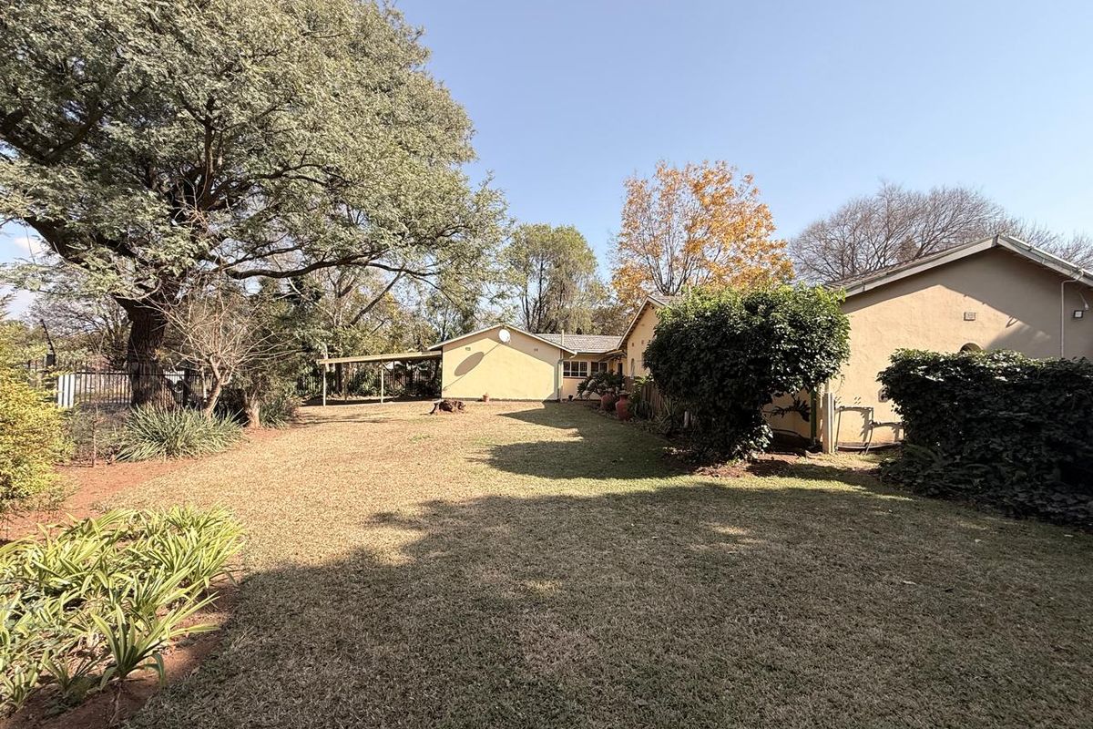 A spacious rear garden featuring a large lawn, mature trees, and a covered patio area attached to the main house structure.