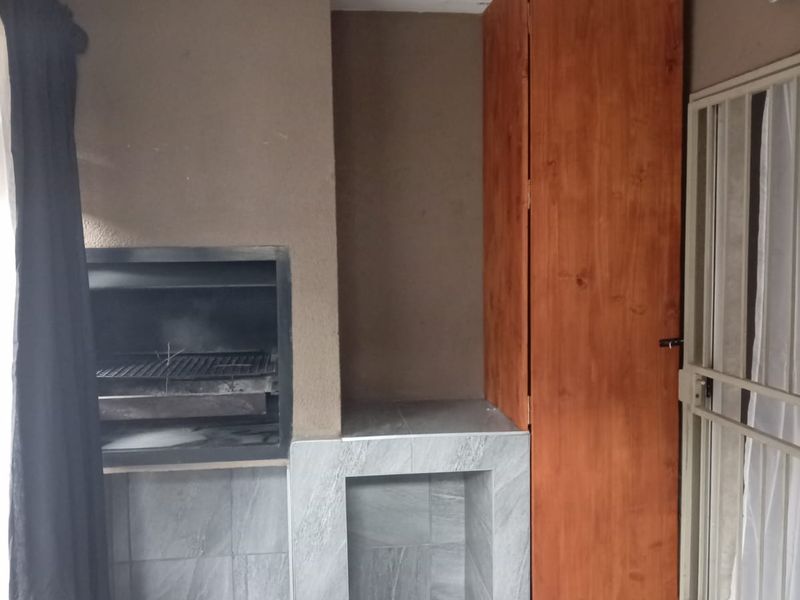 Covered patio area featuring a built-in braai, tiled flooring, and a tall wooden storage cupboard, providing a functional outdoor entertainment space.