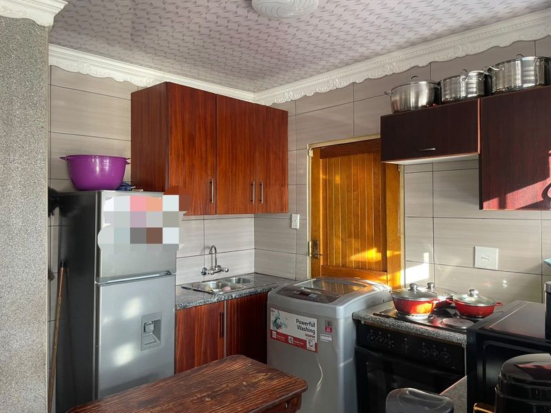 A functional kitchen featuring wood-finish cabinetry, tiled walls, a stainless steel sink, and a freestanding stove with integrated extractor fan.
