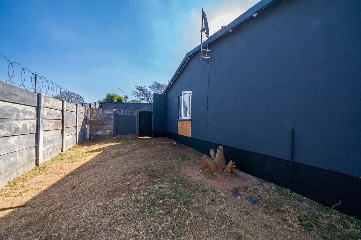 Rear exterior view of the property featuring a dark-painted facade, precast concrete boundary wall with security razor wire, and a small garden area.