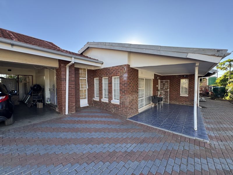 Front exterior view of a brick house featuring a paved driveway, covered carport, and a tiled patio area with security gates.