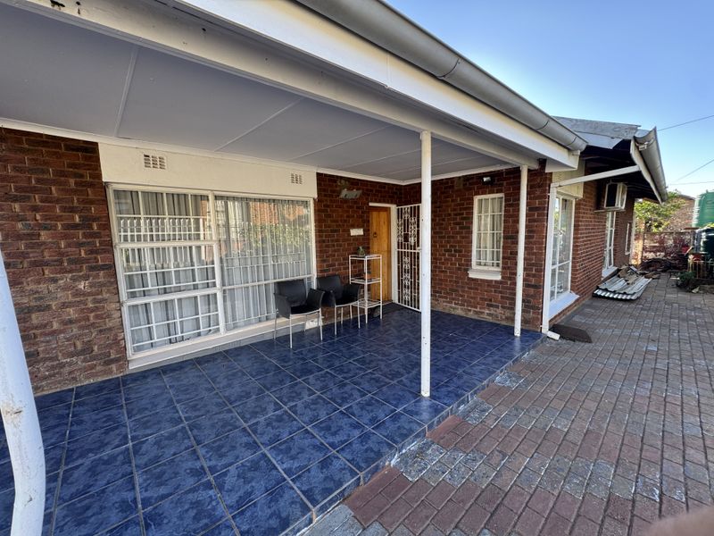 A covered patio area featuring tiled flooring, brick exterior walls, and security fixtures, providing a functional outdoor space adjacent to the main house entrance.
