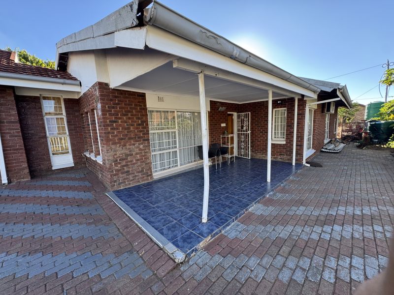 A covered patio area featuring tiled flooring and brick exterior walls, adjacent to a paved driveway and side garden area.