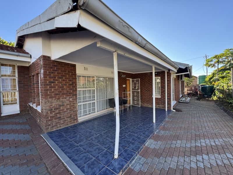 Rear exterior view of a brick house featuring a tiled patio area, paved surroundings, and metal support columns requiring maintenance.