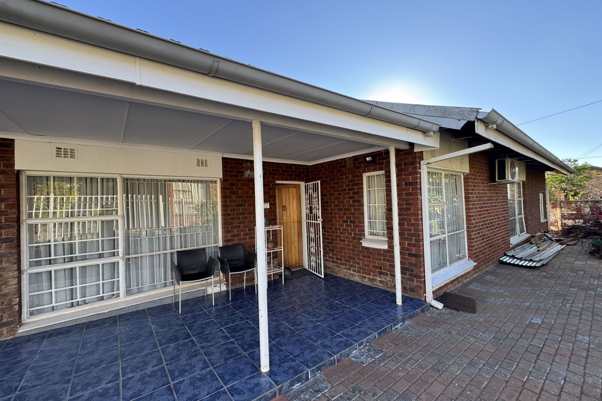 A rear exterior view of a brick house featuring a covered tiled patio, security gates, and paved outdoor area.