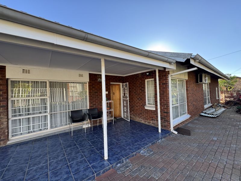 A rear exterior view of a brick house featuring a covered tiled patio, security gates, and paved outdoor area.