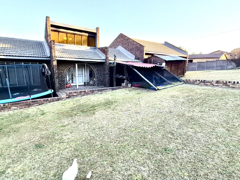 Rear view of the property featuring a brick facade, tiled roof, and a spacious backyard with a retaining wall and covered patio area.