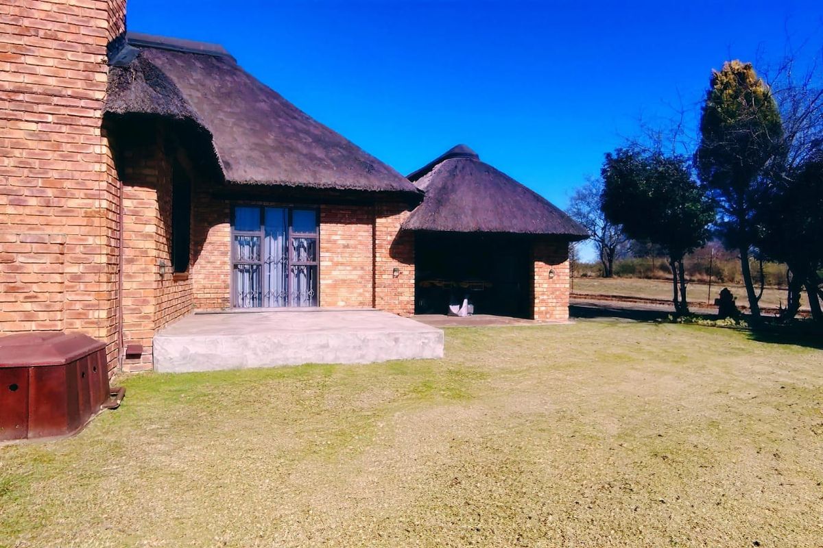 Rear view of a face brick house featuring a traditional thatch roof, a concrete patio, and an open-sided outdoor structure set in a garden.