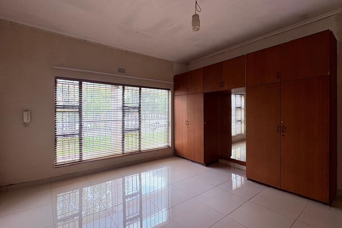 A spacious, empty bedroom featuring large built-in wooden cupboards, polished tiled flooring, and large windows with fitted venetian blinds.