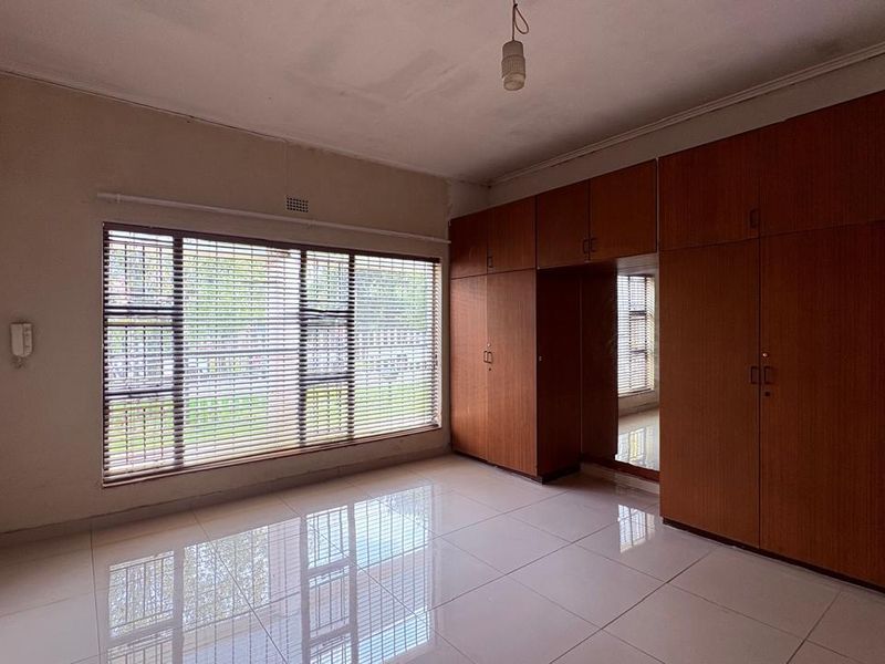 A spacious, empty bedroom featuring large built-in wooden cupboards, polished tiled flooring, and large windows with fitted venetian blinds.