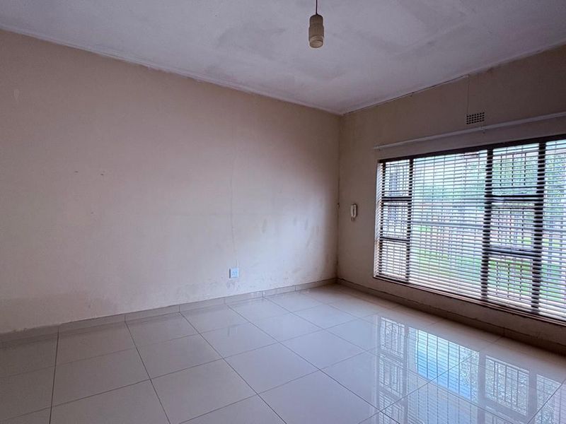 An empty living room featuring tiled flooring and large windows with venetian blinds, requiring minor cosmetic updates to the walls and ceiling.