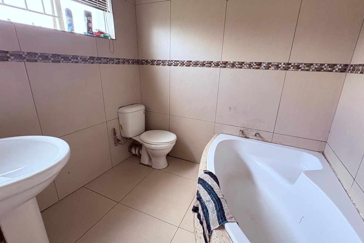 A functional bathroom featuring a corner bathtub, pedestal basin, and toilet, finished with neutral wall and floor tiles and a decorative mosaic border.