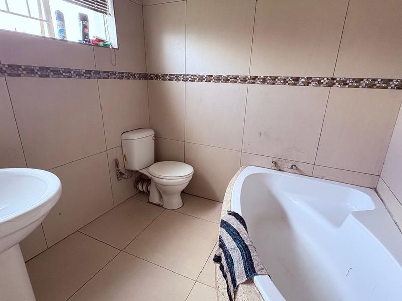 A functional bathroom featuring a corner bathtub, pedestal basin, and toilet, finished with neutral wall and floor tiles and a decorative mosaic border.