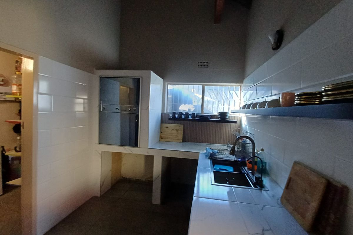 Kitchen featuring stone-look countertops, built-in oven, double sink, and white tiled splashbacks with floating shelving under a high vaulted ceiling.