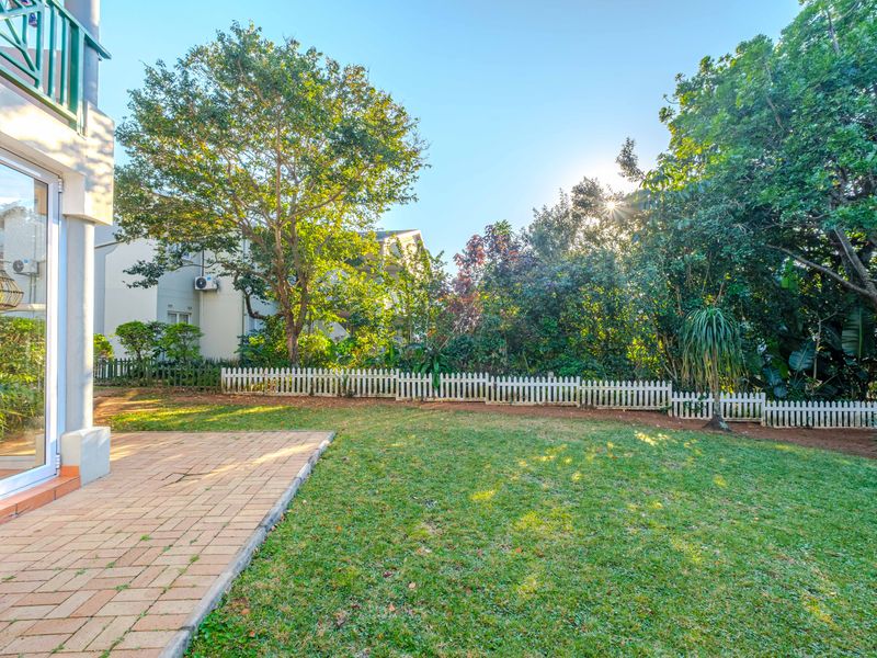 A well-maintained private garden featuring a paved patio area, lush lawn, and a white picket fence bordered by mature trees.