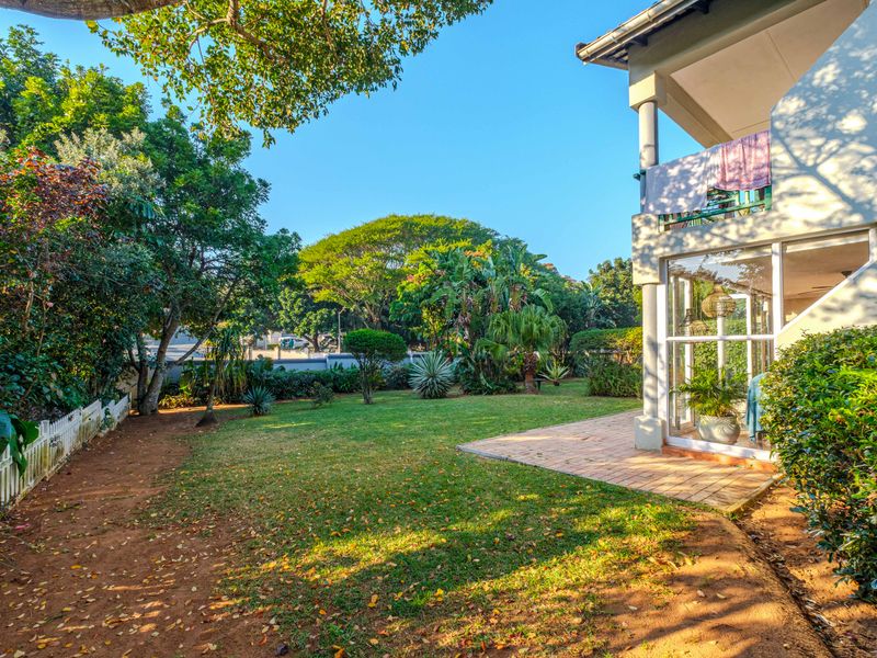 A spacious rear garden featuring a paved patio area, a balcony above, and a well-maintained lawn surrounded by mature trees and a picket fence.