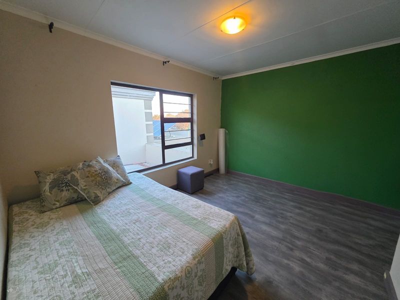 A bedroom featuring laminate flooring, a large aluminium window, and a bold green accent wall, offering a functional space with neutral base tones.