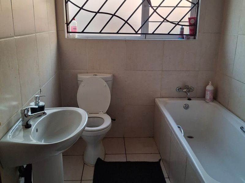 A functional bathroom featuring a pedestal basin, standard toilet, and bathtub with full-height wall tiling and a window with security bars.