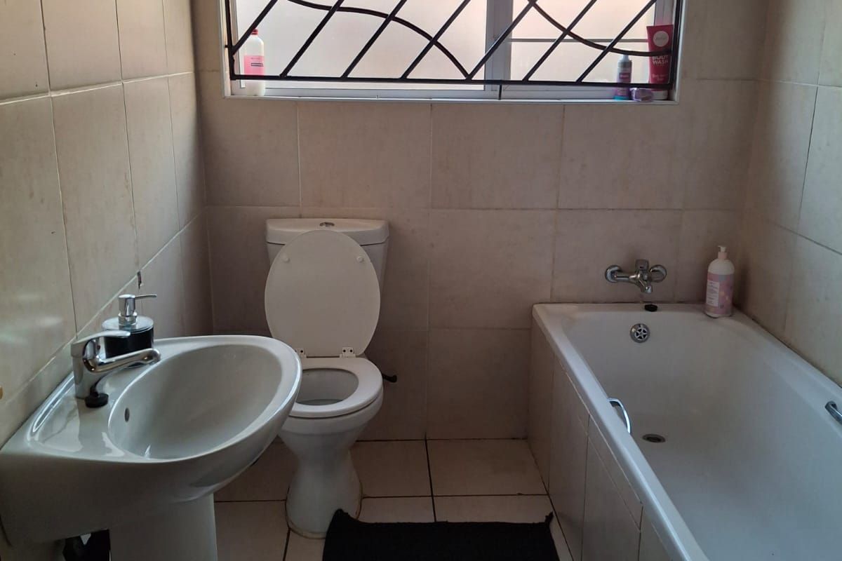 A functional bathroom featuring a pedestal basin, standard toilet, and bathtub with full-height wall tiling and a window with security bars.