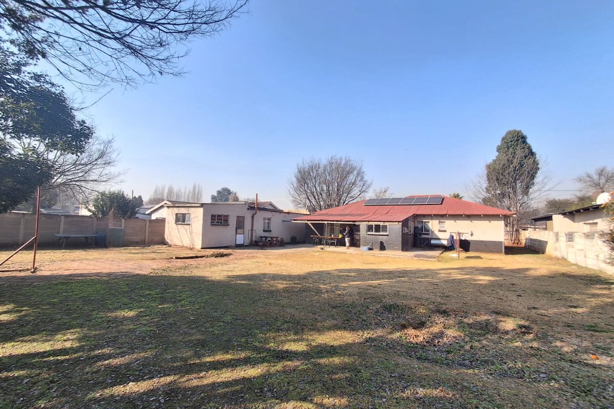 Rear view of the property featuring a large garden, concrete patio area, and a house with a corrugated metal roof and installed solar panels.