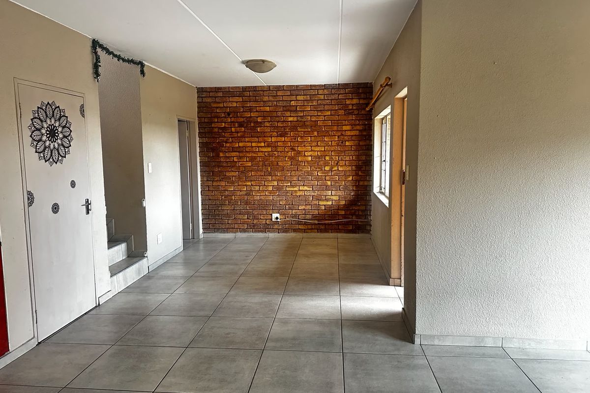 An open-plan living area featuring a prominent exposed brick wall, neutral ceramic floor tiles, and direct access to a staircase.