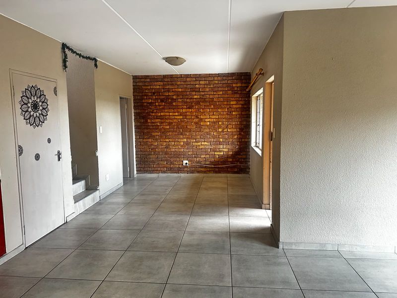 An open-plan living area featuring a prominent exposed brick wall, neutral ceramic floor tiles, and direct access to a staircase.