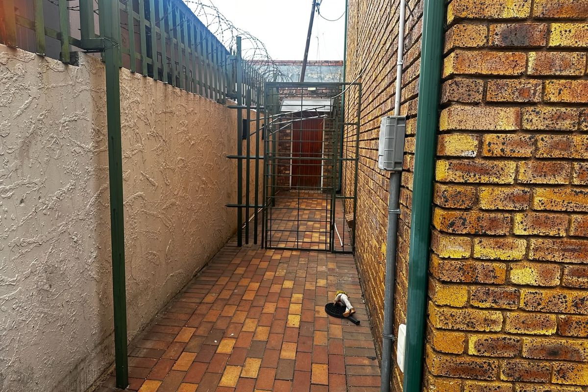 A narrow side passage featuring brick paving, enclosed by a perimeter wall with security fencing and a metal gate.
