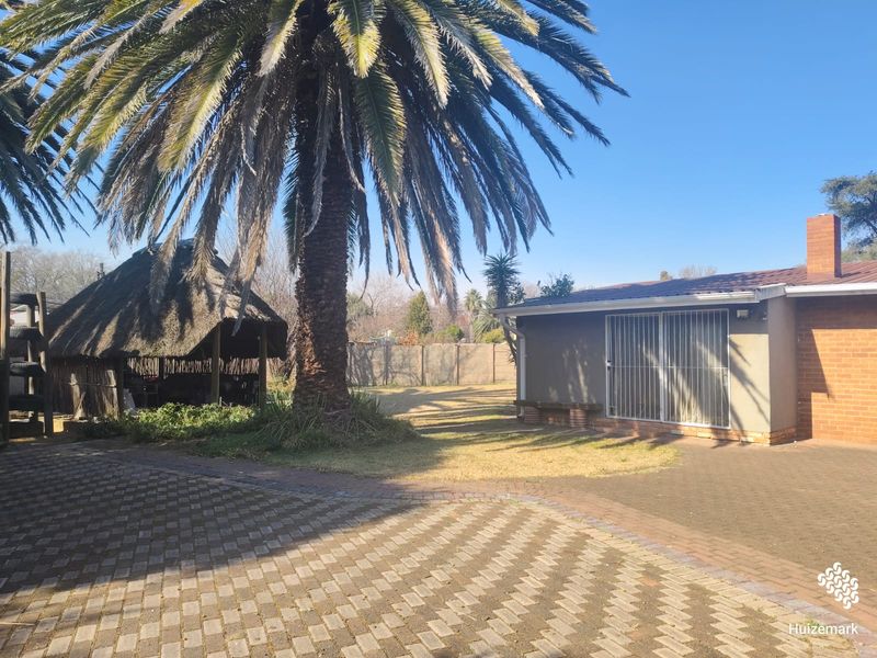 Rear garden area featuring a paved driveway, a traditional thatched lapa, and a brick-built home with security gates.