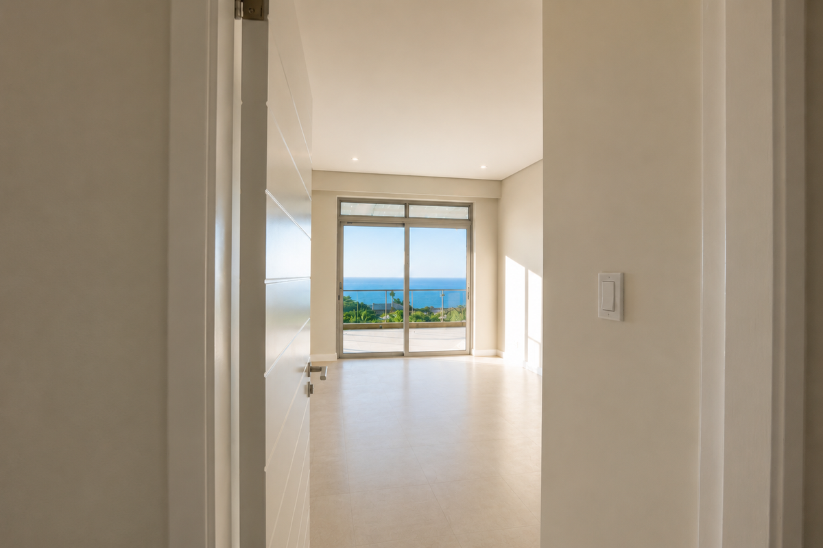 A bright, empty bedroom featuring neutral tiled flooring, recessed lighting, and a large sliding glass door leading to a balcony with ocean views.