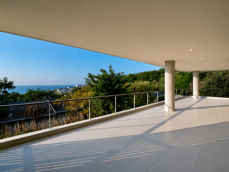 A spacious, modern covered balcony featuring clean tiled flooring, structural columns, and a sleek metal railing with expansive coastal and landscape views.