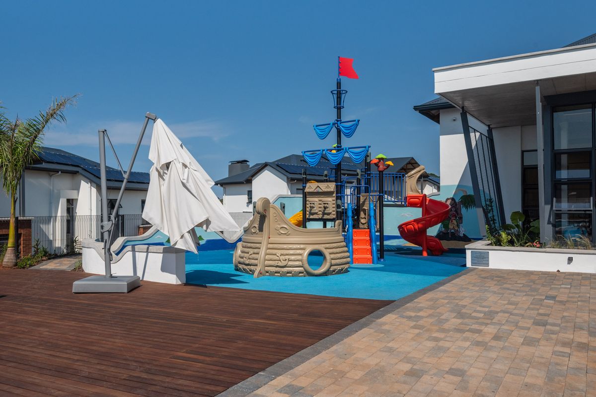 Outdoor communal area featuring a children's playground with rubberized flooring, timber decking, and paved pathways adjacent to the apartment building.