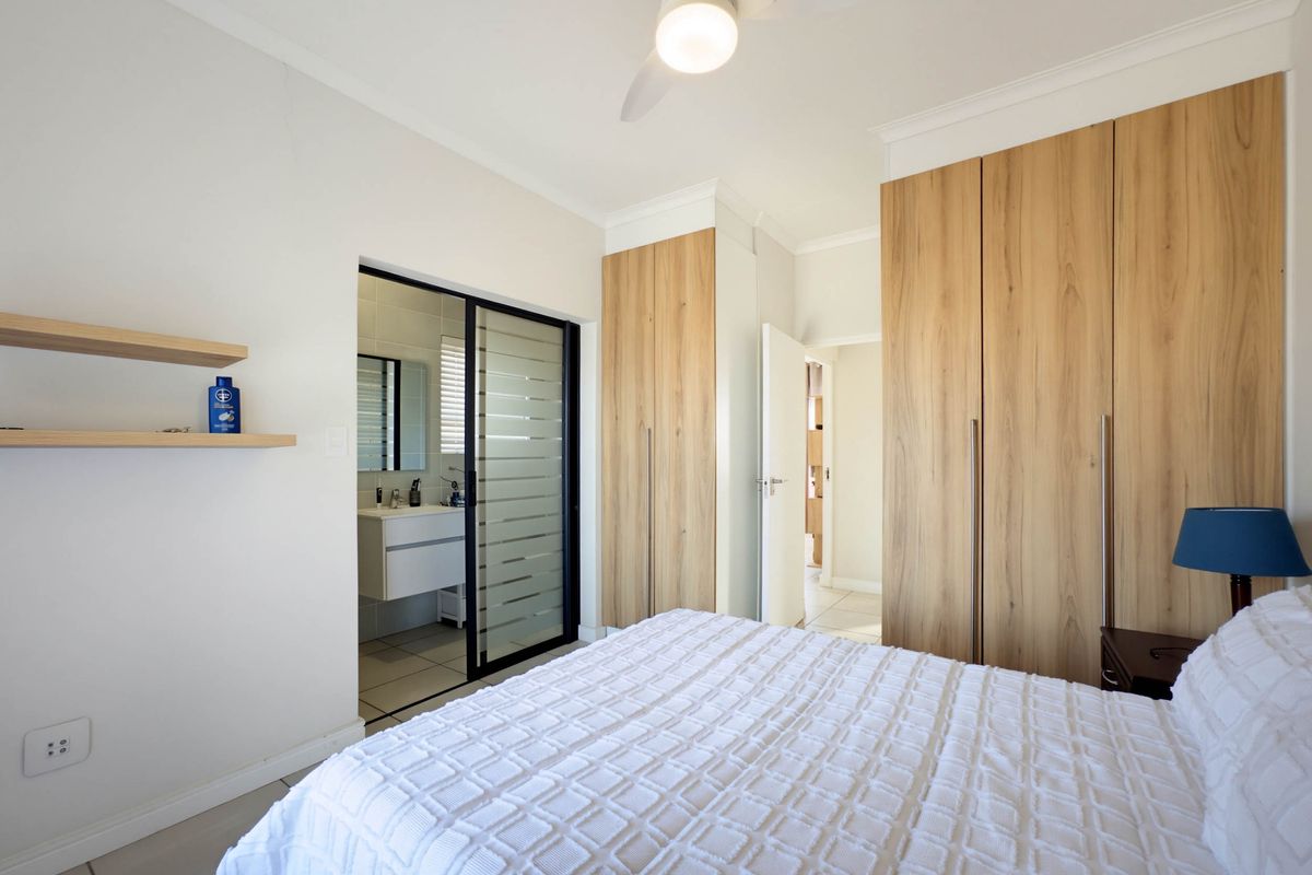 A well-maintained bedroom featuring built-in wooden cupboards, a ceiling fan, and an en-suite bathroom accessed via a modern sliding glass door.