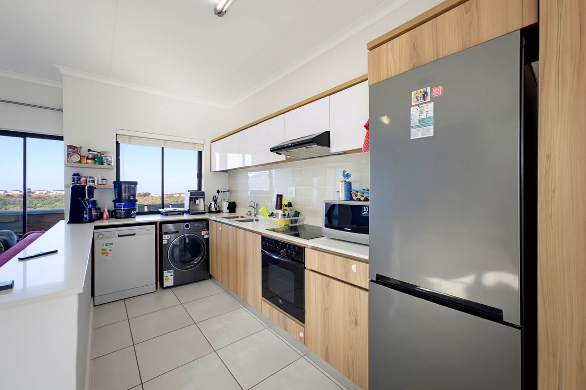Modern kitchen featuring light wood-grain cabinetry, white upper cabinets, tiled backsplash, integrated appliances, and a breakfast bar overlooking the living area.