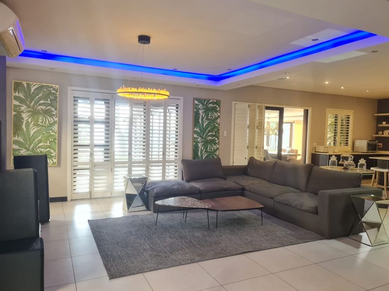 Spacious open-plan living area featuring modern recessed ceiling lighting with blue LED accents, tiled floors, and plantation shutters leading to an outdoor space.